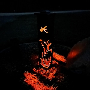 May include: A black metal lantern with a dragonfly and floral design. The lantern is lit and casting a warm orange glow.