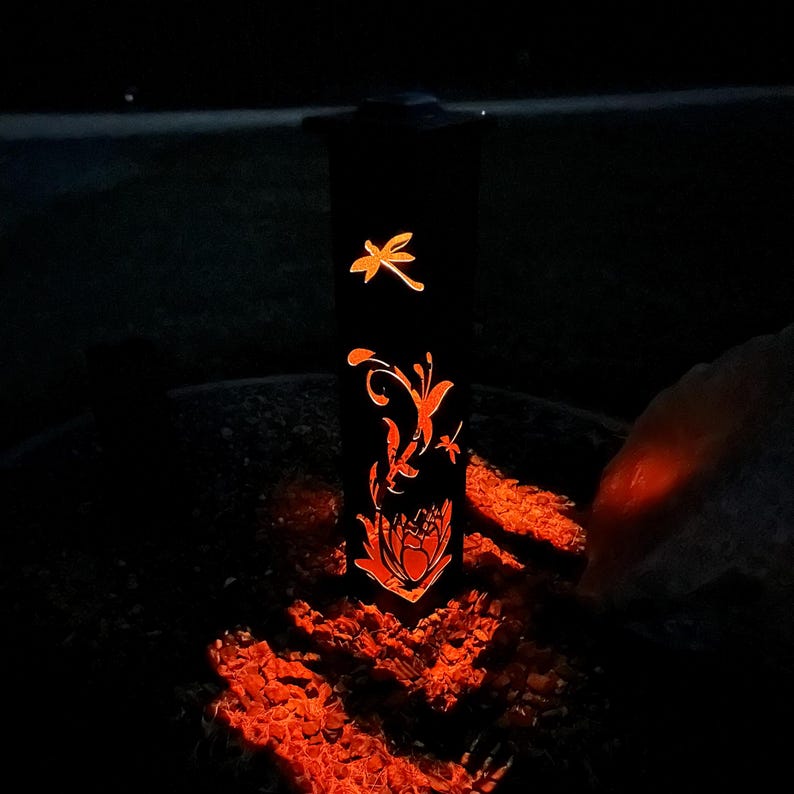 May include: A black metal lantern with a dragonfly and floral design. The lantern is lit and casting a warm orange glow.