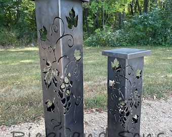 May include: Two metal planters with a grapevine design. The planters are different sizes and are made of a dark metal. The larger planter is on the left and the smaller planter is on the right. The planters are sitting on a gravel surface.