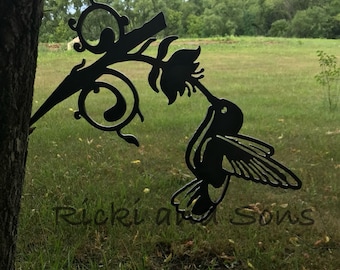 May include: Black metal hummingbird silhouette with a flower attached to a branch. The hummingbird is in flight and has its beak in the flower. The silhouette is attached to a tree trunk. The text "Ricki and Sons" is visible in the bottom right corner of the image.