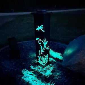 May include: A black metal garden light with a solar panel on top. The light features a cut-out design of a dragonfly, butterfly, and flower. The light is illuminated with a bright blue glow.