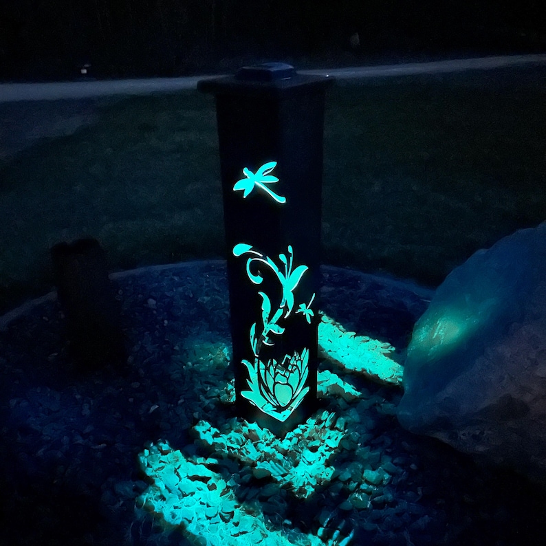 May include: A black metal garden light with a solar panel on top. The light features a cut-out design of a dragonfly, butterfly, and flower. The light is illuminated with a bright blue glow.