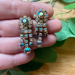 May include: A pair of vintage gold-toned earrings with iridescent, teardrop-shaped crystals. Each earring features a floral-shaped top with a large central stone and dangling crystal accents, creating a glamorous, eye-catching design.