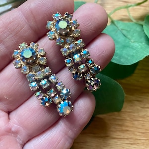 May include: A pair of vintage gold-toned earrings with iridescent, multi-colored rhinestones. The earrings feature a floral design at the top and a cascading, chandelier-style drop. The rhinestones reflect shades of blue, green, and purple.