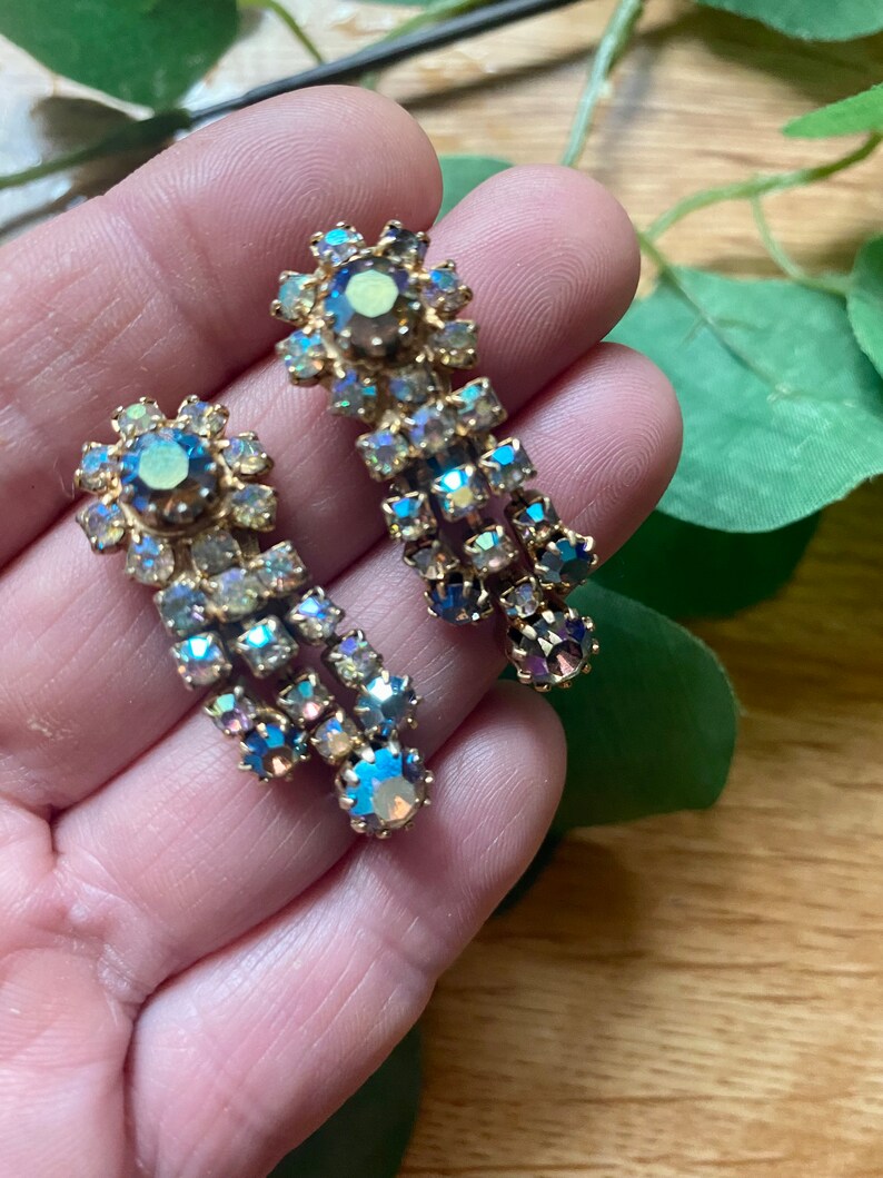 May include: A pair of vintage gold-toned earrings with iridescent, multi-colored rhinestones. The earrings feature a floral design at the top and a cascading, chandelier-style drop. The rhinestones reflect shades of blue, green, and purple.
