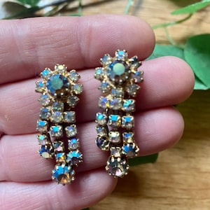 May include: A pair of vintage gold-toned earrings with iridescent, rectangular and round crystals. The earrings have a floral design at the top and a cascading, dangling crystal arrangement. The crystals reflect shades of blue, purple, and yellow.