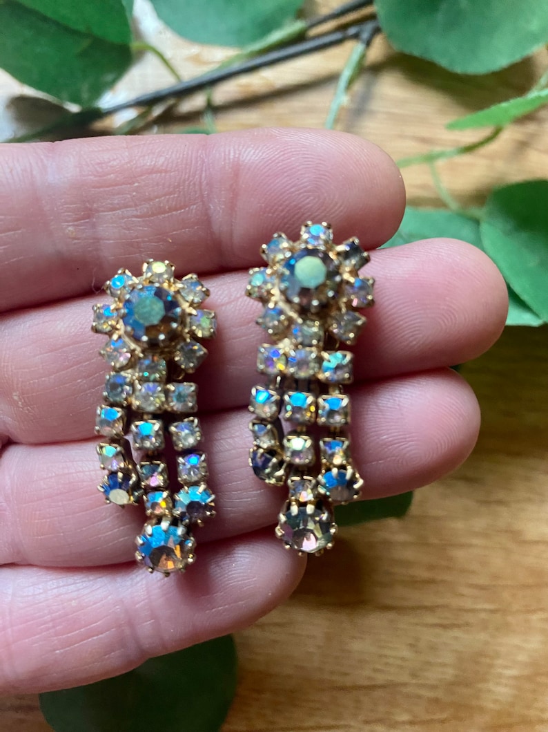 May include: A pair of vintage gold-toned earrings with iridescent, rectangular and round crystals. The earrings have a floral design at the top and a cascading, dangling crystal arrangement. The crystals reflect shades of blue, purple, and yellow.