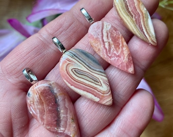 May include: Four teardrop-shaped gemstone pendants with silver-colored bails. The pendants display a variety of pink, cream, and brown banded patterns. The gemstones are held in a hand, with a blurred purple flower in the background.