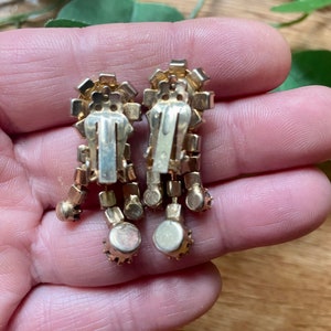 May include: A pair of vintage clip-on earrings. The earrings have a gold-tone metal setting with clear and amber-colored rhinestones. The design features a rectangular top with dangling, round-shaped accents.