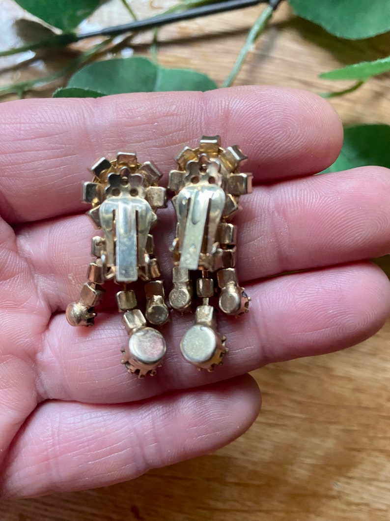 May include: A pair of vintage clip-on earrings. The earrings have a gold-tone metal setting with clear and amber-colored rhinestones. The design features a rectangular top with dangling, round-shaped accents.