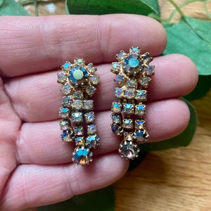 May include: A pair of vintage gold-toned earrings with iridescent, teardrop-shaped rhinestones. The earrings feature a floral design at the top and a cascading arrangement of rectangular and round stones, creating a glamorous, dangling effect. These earrings are perfect for special occasions.