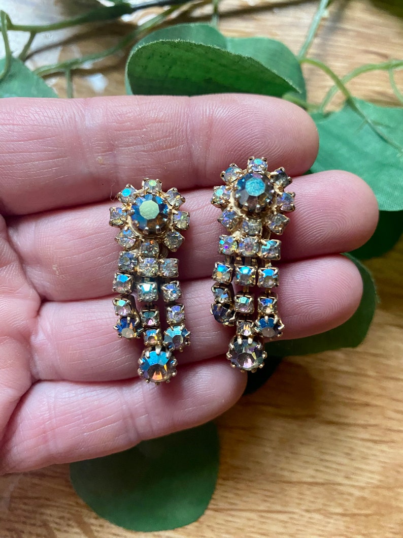 May include: A pair of vintage gold-toned earrings with iridescent, teardrop-shaped rhinestones. The earrings feature a floral design at the top and a cascading arrangement of rectangular and round stones, creating a glamorous, dangling effect. These earrings are perfect for special occasions.