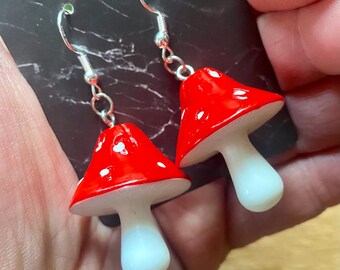May include: A pair of mushroom earrings. Each earring features a red cap with white spots and a white stem. The earrings are suspended from silver-colored fish hook ear wires.