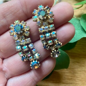 May include: A pair of vintage gold-toned earrings with iridescent, multi-colored rhinestones. The earrings feature a floral design at the top and a cascading arrangement of stones. The stones reflect shades of blue, green, and purple.