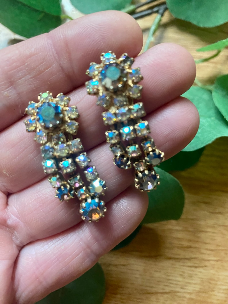 May include: A pair of vintage gold-toned earrings with iridescent, multi-colored rhinestones. The earrings feature a floral design at the top and a cascading arrangement of stones. The stones reflect shades of blue, green, and purple.