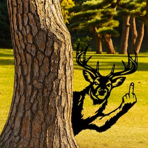 Peut inclure: Silhouette noire d'un cerf en métal, apparaissant derrière un arbre et montrant le majeur. Le cerf a de grands bois et se détache sur un fond d'herbe verte et d'arbres.