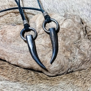 May include: Two pendant necklaces with dark metal horn-shaped charms. Each charm is attached to a silver ring and a black cord. The pendants rest on a piece of weathered wood, with a fur-like background.
