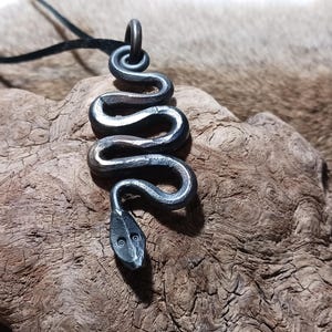 May include: A handcrafted, metal snake pendant on a black cord. The snake is coiled with a detailed head and body, and is displayed on a textured, brown surface. The pendant is a unique piece of jewelry.