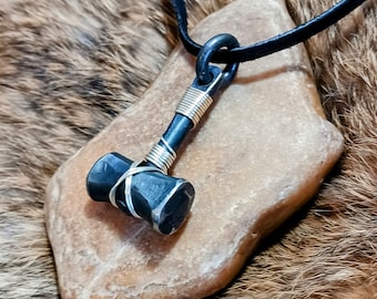 May include: A handcrafted pendant necklace with a miniature hammer design. The hammer is made of dark metal, wrapped with silver and gold-coloured wire, and hangs from a black cord. The pendant rests on a light brown stone.