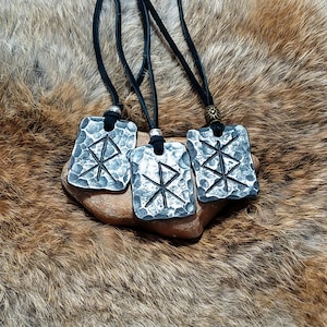 May include: Four silver pendants with a black leather cord, each featuring a hammered metal design with a Norse rune symbol. The pendants are displayed on a brown fur surface.