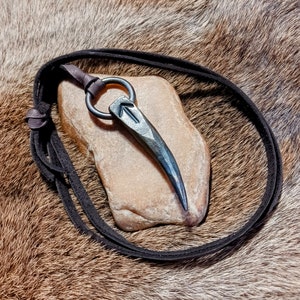 May include: A black leather cord necklace with a silver pendant shaped like a sharp, pointed arrowhead. The pendant is resting on a light brown rock.
