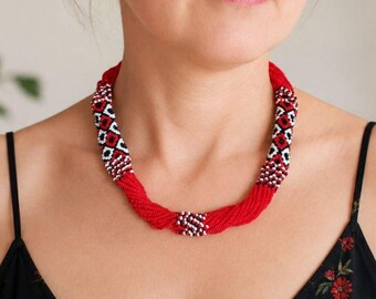 May include: A multi-strand beaded necklace featuring a vibrant red, black, and white geometric pattern. The design consists of numerous small beads woven into a thick, rounded cord structure that sits close to the neck in a circular shape.