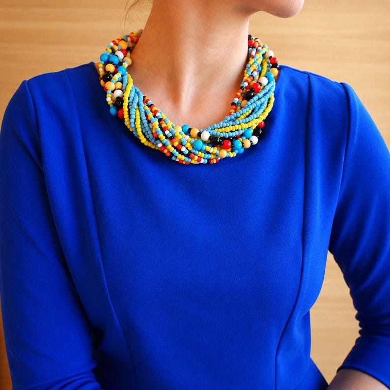 May include: A multi-strand statement necklace featuring numerous small beads in vibrant shades of yellow, turquoise blue, red, black, and white. The beads are arranged in a twisted, layered design that sits closely around the neck, creating a dense and colorful circular pattern of individual spherical elements.