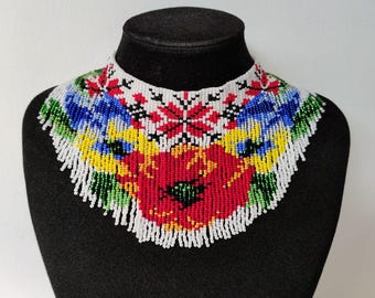 May include: A colorful beaded necklace with a floral design. The necklace features red, yellow, blue, green, and white beads arranged in a pattern of flowers and leaves. The necklace is on a black mannequin.