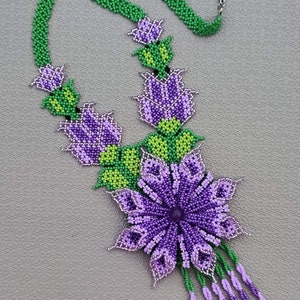 Könnte beinhalten: Eine Perlenkette mit einem Anhänger in Form einer violetten Blume und grünen Blättern. Die Blume hat eine violette Mitte und die Blätter sind grün. Die Kette besteht aus kleinen Perlen und hat einen silbernen Verschluss.
