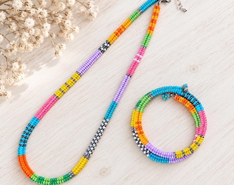 May include: A beaded necklace and matching bracelet set featuring vibrant, multi-colored glass seed beads arranged in repeating segments of orange, blue, green, yellow, pink, and purple, accented with small silver-toned metal spacer beads.