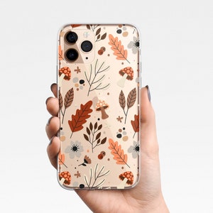 Puede incluir: Funda de tel&eacute;fono transparente con un dise&ntilde;o de tem&aacute;tica oto&ntilde;al. El dise&ntilde;o incluye hojas marrones y naranjas, setas y peque&ntilde;as flores blancas sobre un fondo beige. La funda se sostiene en una mano.