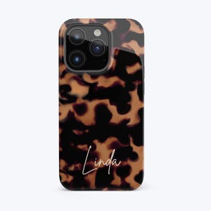 Puede incluir: Funda de teléfono brillante con un diseño de carey en tonos marrones y negros. El nombre "Linda" está escrito en cursiva en la parte inferior de la funda. La funda tiene un recorte para la cámara.