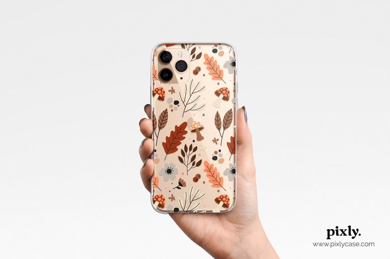 Puede incluir: Funda de tel&eacute;fono transparente con un dise&ntilde;o de tem&aacute;tica oto&ntilde;al. El dise&ntilde;o incluye hojas marrones y naranjas, setas y peque&ntilde;as flores blancas sobre un fondo beige. La funda se sostiene en una mano.