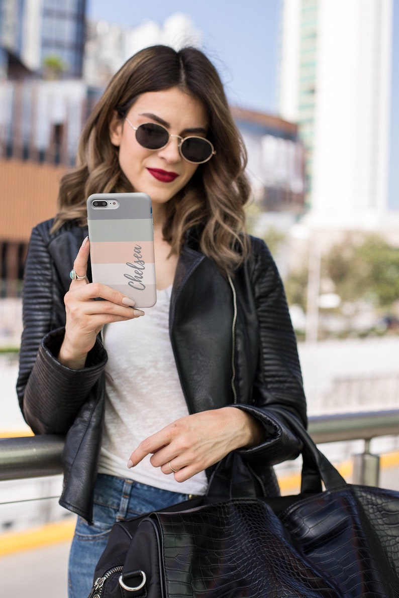 May include: A woman wearing a black leather jacket, sunglasses, and a white t-shirt is holding a phone case with the name "Chelsea" printed on it. The phone case has a gray, pink, and white striped design.
