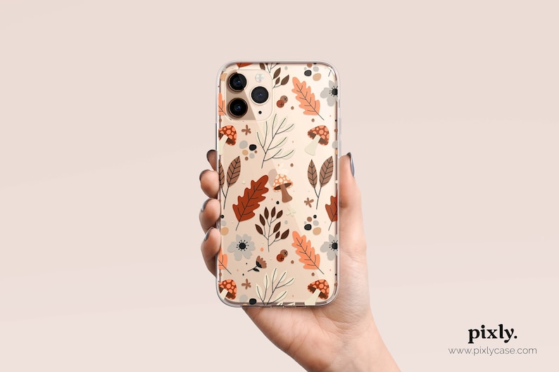 Puede incluir: Funda transparente para tel&eacute;fono con un dise&ntilde;o oto&ntilde;al. El dise&ntilde;o incluye hojas, setas y flores marrones y naranjas sobre un fondo beige. La funda se muestra en una mano, mostrando el estampado detallado.