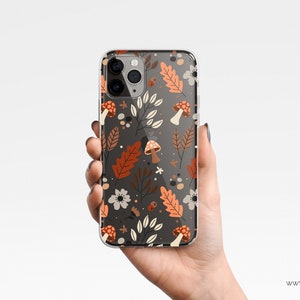 Puede incluir: Funda transparente para tel&eacute;fono con un dise&ntilde;o oto&ntilde;al. El dise&ntilde;o incluye hojas naranjas y marrones, flores blancas y setas sobre un fondo gris oscuro. La funda est&aacute; sujeta en una mano.