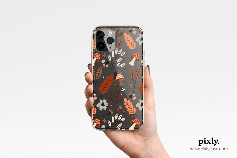 Puede incluir: Funda transparente para tel&eacute;fono con un dise&ntilde;o oto&ntilde;al. El dise&ntilde;o incluye hojas naranjas y marrones, flores blancas y setas sobre un fondo gris oscuro. La funda est&aacute; sujeta en una mano.