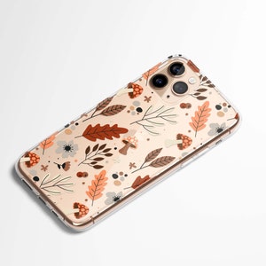 Puede incluir: Funda transparente para tel&eacute;fono con dise&ntilde;o oto&ntilde;al. La funda presenta un patr&oacute;n de hojas marrones y naranjas, setas y flores sobre un fondo beige. La funda del tel&eacute;fono est&aacute; en &aacute;ngulo, mostrando las lentes de la c&aacute;mara.