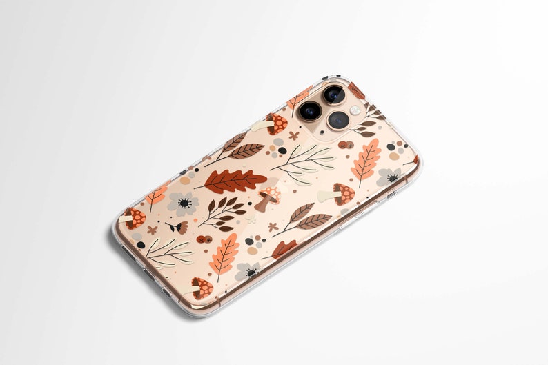 Puede incluir: Funda transparente para tel&eacute;fono con dise&ntilde;o oto&ntilde;al. La funda presenta un patr&oacute;n de hojas marrones y naranjas, setas y flores sobre un fondo beige. La funda del tel&eacute;fono est&aacute; en &aacute;ngulo, mostrando las lentes de la c&aacute;mara.