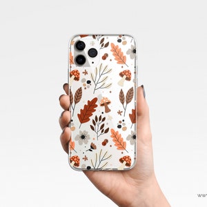 Puede incluir: Funda transparente para tel&eacute;fono con un patr&oacute;n repetido de hojas de oto&ntilde;o, setas y flores en tonos marr&oacute;n, naranja y crema. La funda se sostiene en una mano sobre un fondo blanco. La funda tiene un acabado brillante.