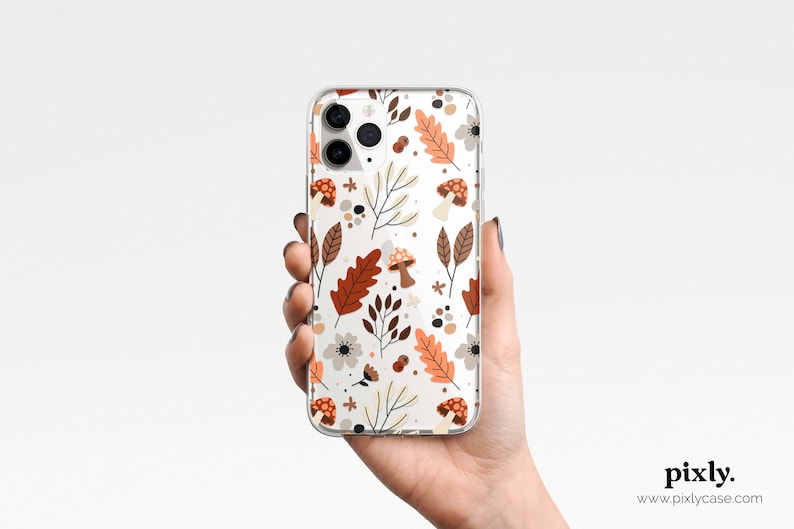 Puede incluir: Funda transparente para tel&eacute;fono con un patr&oacute;n repetido de hojas de oto&ntilde;o, setas y flores en tonos marr&oacute;n, naranja y crema. La funda se sostiene en una mano sobre un fondo blanco. La funda tiene un acabado brillante.