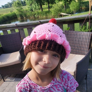 Puede incluir: Una niña pequeña lleva una gorra de cupcake de ganchillo rosa y marrón con una cereza roja en la parte superior. Lleva una camisa de estampado rosa y blanco con un diseño de signo de paz.