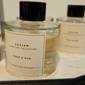 May include: A clear glass bottle with a black lid and a label that reads "Zavian Signature Collection Rose & Oud Luxury Reed Diffuser". The bottle is sitting on a white surface with black reed diffusers.