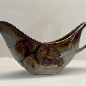 May include: A ceramic gravy boat featuring a glossy glaze with a blend of earthy brown, tan, and cream tones. The vessel has a curved, elongated silhouette with a pointed pouring spout on one side and a looped handle on the opposite side.