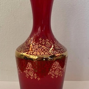 May include: A tall, ruby red glass vase featuring a flared rim and a cylindrical body. The vase is decorated with intricate gold filigree patterns, including floral motifs and scrollwork, along with a thick gold band around the centre and rim.