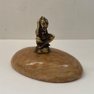 May include: A polished, oval-shaped tan and cream banded stone base topped with a small, seated brass figure. The figure has a pointed head, defined facial features, and is depicted in a cross-legged position, securely mounted to the centre of the stone.