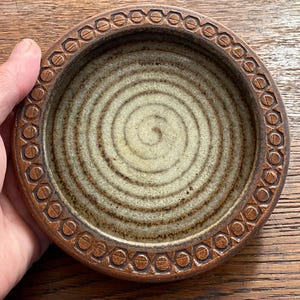 May include: A round ceramic bowl with a light beige interior featuring a dark brown spiral pattern. The thick rim is a deep brown colour and is decorated with a repeating geometric pattern of small circles and vertical lines around the edge.