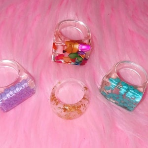 Puede incluir: Cuatro anillos de resina transparente con diferentes inclusiones coloridas. Un anillo tiene cuentas moradas, otro tiene copos dorados, un tercero tiene cuentas turquesas y el último tiene una mezcla de piedras coloridas.