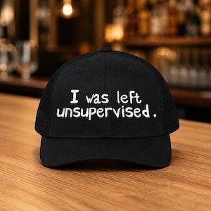 May include: Black trucker cap featuring the white text "I was left unsupervised." The cap has a mesh back and a curved peak. The hat is displayed on a wooden surface.