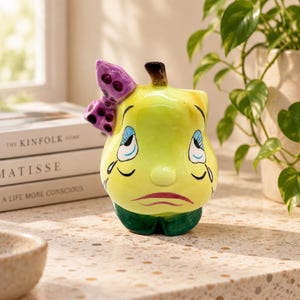 May include: A ceramic pear-shaped figurine with a glossy yellow finish, a dark green base, and a brown stem. The pear features a painted face with closed eyes, blue eyelids, and a sad mouth, accented by a purple bow with black polka dots.